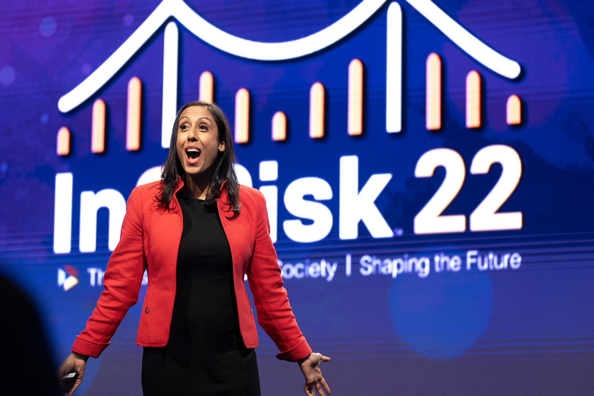 A passionate speaker gestures animatedly in front of a presentation with the words "in2risk 22" on the screen, conveying excitement or a pivotal moment in her speech.
