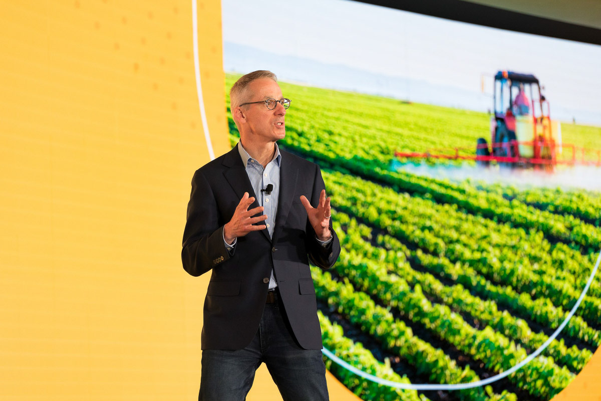 A speaker presenting at a conference with a large agricultural image in the background.