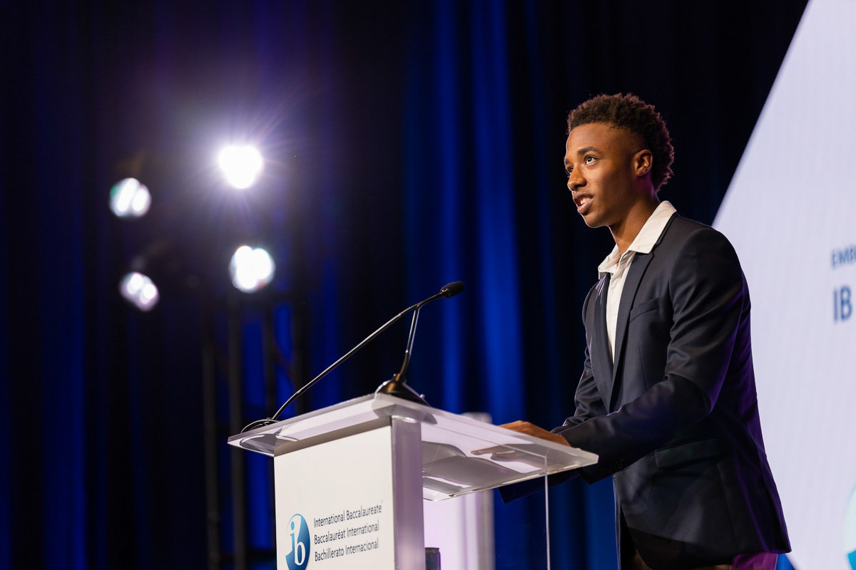 A young man confidently delivers a speech at a podium with conference banners in the background and stage lights illuminating the scene.